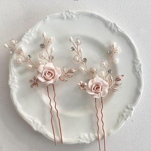 May include: Two rose gold hair pins with delicate floral designs. Each pin features a pink rose, pearl accents, and crystal embellishments.