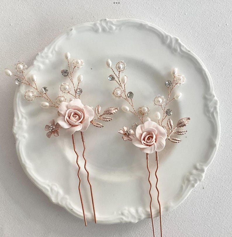May include: Two rose gold hair pins with delicate floral designs. Each pin features a pink rose, pearl accents, and crystal embellishments.