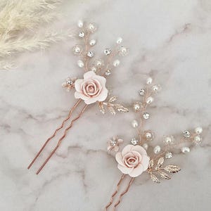May include: Two rose gold hair pins with delicate floral designs. Each pin features a pink rose with pearl and crystal accents. The pins are perfect for adding a touch of elegance to any hairstyle.