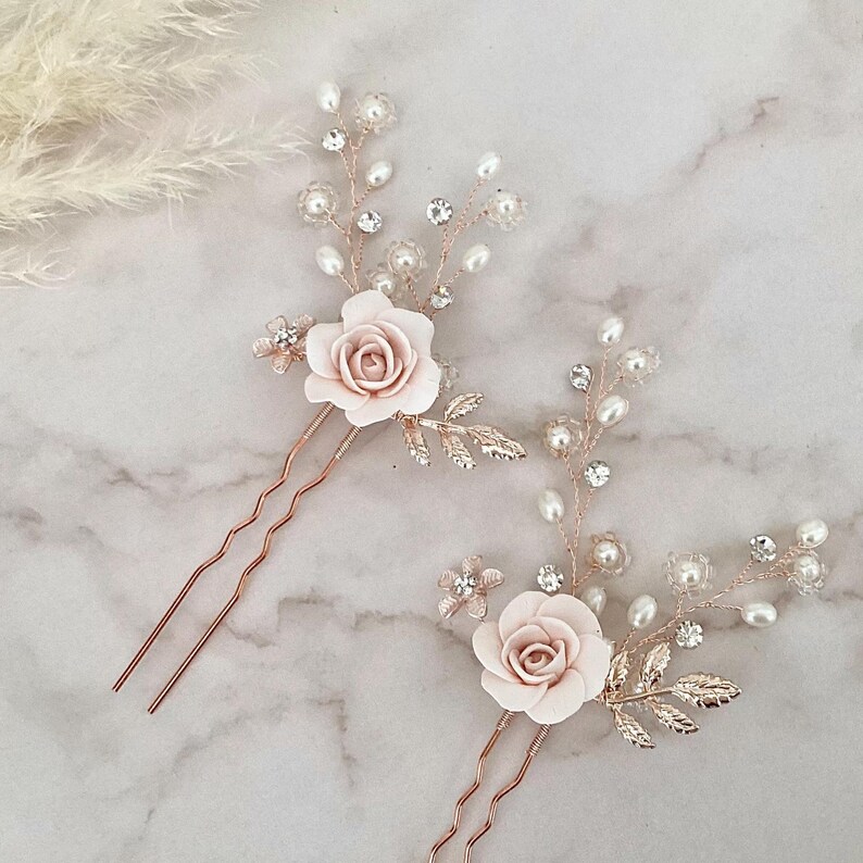 May include: Two rose gold hair pins with delicate floral designs. Each pin features a pink rose with pearl and crystal accents. The pins are perfect for adding a touch of elegance to any hairstyle.