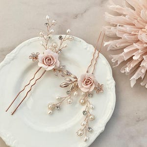 May include: Two gold hair pins with delicate floral designs. Each pin features a blush pink flower with pearl and crystal accents.