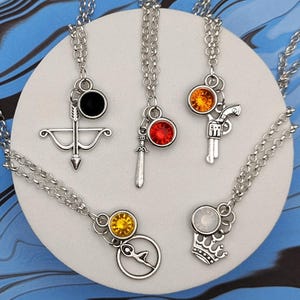 Puede incluir: Una colección de collares plateados con varios diseños de colgantes, incluyendo un arco y flecha, una espada, una pistola, una brújula y una corona. Cada colgante presenta una gema de color. Los collares se exhiben sobre una superficie circular blanca contra un fondo marmoleado azul y negro.