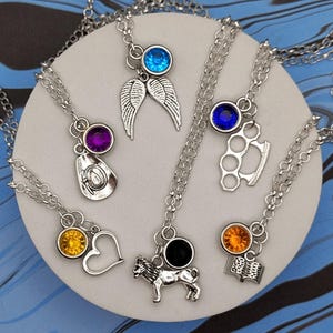 Puede incluir: Una colección de collares plateados con varios diseños de colgantes, incluyendo un corazón, alas, un sombrero de vaquero, un perro y unos nudillos de latón. Cada collar presenta una gema de color. Los collares se exhiben sobre una superficie circular blanca contra un fondo azul y negro veteado.