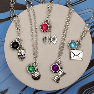 May include: A collection of silver-toned necklaces with various charms, including a skull, an axe, a basketball, an hourglass, and an envelope. Each necklace features a small, colorful gemstone. The necklaces are displayed on a white circular surface against a blue and black marbled background.