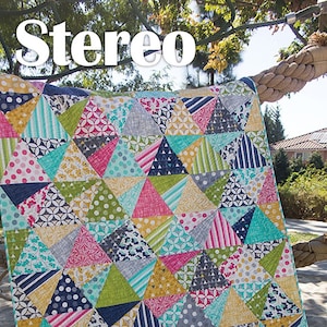 May include: A colorful quilt with a geometric pattern of triangles in shades of blue, green, pink, yellow, and white. The quilt is made of various fabrics with different patterns, including polka dots, stripes, and floral designs. The quilt is hanging on a rope and the text "Stereo" and "JAYBIRD QUILTS" is visible.