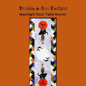 May include: A Halloween-themed table runner featuring a black cat, a pumpkin, and a small owl perched on top of the pumpkin. The background is a gray and white moon with bats flying around it. The table runner is made of fabric with a patchwork design in black, white, green, and purple.