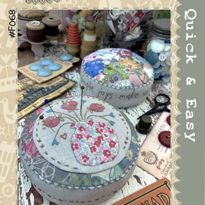 May include: Two handmade pincushions, one with a floral pattern and the other with a vase and flowers embroidered on it. The pincushions are on a wooden surface with other sewing supplies, including spools of thread, buttons, and a thimble. The text "Simple Joys Pincushions" is at the bottom of the image.