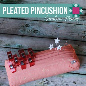 May include: A pink pleated pincushion with a floral pattern, featuring red clips and star-shaped pins. The text "PLEATED PINCUSHION" and "Carolina Moore" are displayed on a teal background. The pincushion is on a wooden surface.