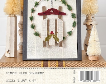May include: A felt winter sled ornament with a red hat and holly leaves. The ornament is framed in a black frame and surrounded by a green wreath. The ornament is 3.25 inches by 5 inches.
