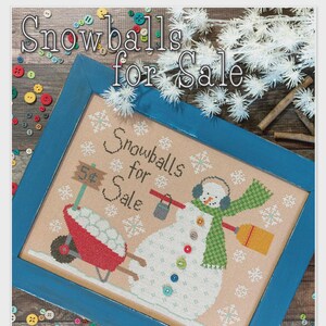 May include: A framed cross-stitch featuring a snowman with a green scarf and earmuffs, holding a broom and a bucket of snowballs. The text "Snowballs for Sale" is displayed, along with snowflakes. The frame is blue, and the background is a light tan color.