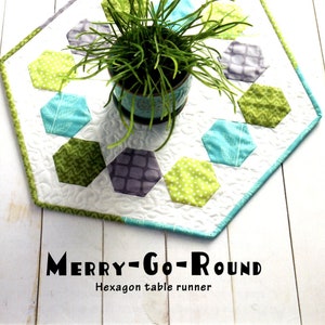 May include: A white hexagon table runner with a green, blue, and gray geometric pattern. The table runner is made of fabric and has a white background. The text "MERRY-GO-ROUND" and "Hexagon table runner" is printed on the bottom of the image.
