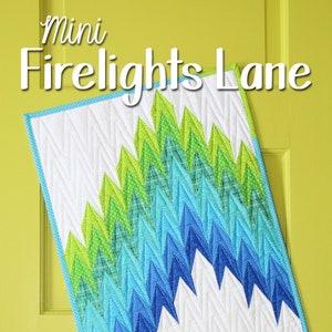 May include: A mini quilt with a "Firelights Lane" title. The quilt features a design of blue and green triangles arranged in a chevron pattern. The quilt is edged with a light blue trim.