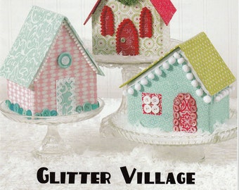 May include: Three miniature houses decorated with colorful paper and fabric. The houses are on white cake stands and have a green, pink, and blue color scheme. The houses are decorated with buttons, fabric scraps, and other embellishments. The image is titled "Glitter Village" and has the text "Atkinson Designs, one of life's simple pleasures, ATK-165".
