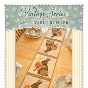 May include: A table runner featuring a patchwork design with brown bunnies holding Easter eggs. The runner is made up of five squares with a brown bunny and egg design on each square. The runner is on a wooden table with a white tablecloth. The text "The Vintage Series April Table Runner" is at the top of the image.