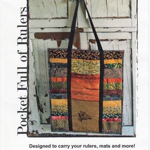 May include: A patchwork fabric tote bag with black straps, designed to carry rulers, mats, and more. The bag is hanging on a white wooden door with the text "Pocket Full of Rulers" and "BELLA NONNA 156 pattern no." printed on the image.