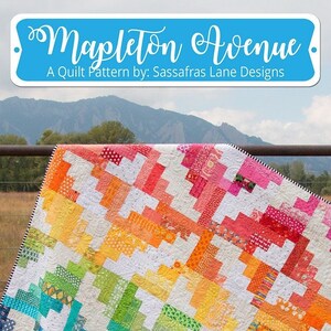 May include: A colorful quilt with a rainbow gradient design, featuring a patchwork pattern of various fabrics. The quilt is draped over a metal fence with a mountain backdrop. The text "Mapleton Avenue" and "A Quilt Pattern by: Sassafras Lane Designs" are visible.