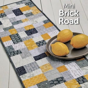 May include: A quilting pattern for a table runner called "Mini Brick Road" by Atkinson Designs. The pattern features a patchwork design with gray, white, and yellow squares. The pattern is shown on a table runner with lemons on a plate.