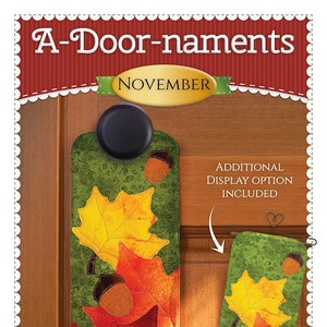 May include: A-Door-naments November door hanger with a green background and fall leaves in shades of red, orange, and yellow with acorns. The door hanger is made of fabric and has an additional display option included.