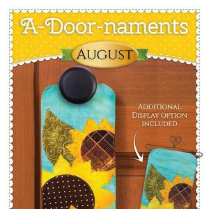 May include: A-Door-naments August door hanger featuring a sunflower design with a blue background. The hanger is made of fabric and has a black knob for hanging. The image also shows an additional display option with a smaller version of the hanger.