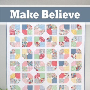 May include: A quilt with a "Make Believe" title, featuring a patchwork design of colorful floral and geometric patterns in shades of pink, blue, green, yellow, and white. The quilt is framed with a light blue border. The quilt is hanging on a white wall with a white table and a potted plant in front of it. A logo for "a bright corner" is in the bottom right corner of the image.