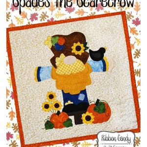May include: A colorful patchwork quilt block featuring a scarecrow with a brown hat, sunflowers, pumpkins, and a black bird. The quilt block is surrounded by a white background with a pattern of orange and brown leaves. The text "Gnome-Mania!! September Spades the Scarecrow" is at the top of the image. The text "Ribbon Candy Quilt Company" is at the bottom of the image.
