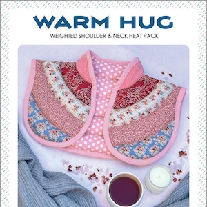 May include: A pink and blue quilted weighted shoulder and neck heat pack with a floral pattern. The heat pack is on a white surface with a gray knitted scarf, a cup of tea, and a candle.