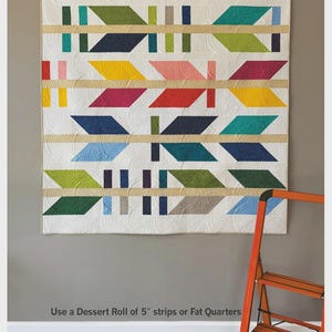 May include: A colorful quilt with an arrow design, featuring various colors like green, blue, yellow, and pink. The quilt is displayed on a wall, with the text "THE ARROW MAKER" at the top. The quilt pattern is by Robin Pickens.