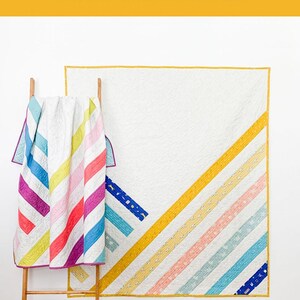 May include: Two quilts with a diagonal pattern of colorful stripes. The quilt on the left is folded and hanging on a wooden ladder. The quilt on the right is hanging on a white wall. The quilts are made with a white background and colorful stripes. The text "MIDPOINT" is printed in white on a yellow background.