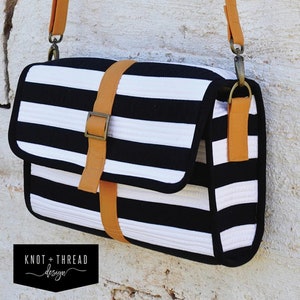 May include: A black and white striped satchel bag with a brown leather strap and buckle. The bag has a top flap with a buckle closure. The bag is labeled "CAMDEN STREET satchel" and "KNOT + THREAD design".