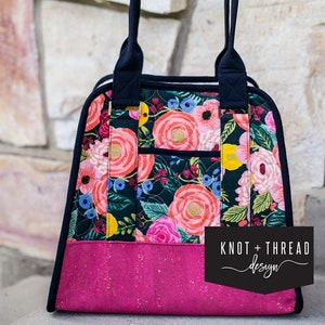 May include: A black and pink floral tote bag with black handles and a black pocket on the front. The bag has the text "Knot + Thread design" on the bottom right corner.