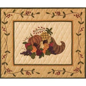 May include: A quilted placemat with a brown, green, and cream color scheme. The center features an embroidered cornucopia overflowing with fall harvest items, including a pumpkin, pineapple, grapes, and apples.