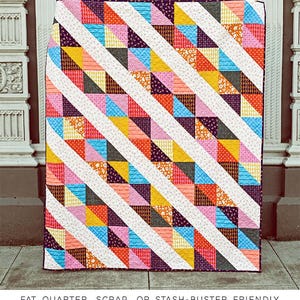 May include: A colorful quilt pattern called "The Daphne Quilt" by Erica Jackman. The quilt is made with a variety of fabrics in shades of pink, blue, yellow, orange, and red. The quilt is made up of triangles and squares. The quilt is displayed on a sidewalk in front of a building. The text "KITCHEN TABLE QUILTING" is at the bottom of the image.
