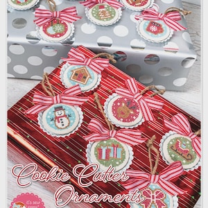 May include: A collection of cross-stitch cookie cutter ornaments with red and white striped bows and twine hangers. Each ornament features a different holiday design, including a snowman, gingerbread house, and Christmas tree. The text "Cookie Cutter Ornaments" is visible.