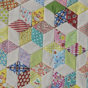 May include: Close-up of a colorful quilt with a geometric pattern. The quilt features a repeating design of three-dimensional cubes, created from various patterned fabrics in shades of yellow, red, green, blue, and pink. The cubes are separated by white fabric with small, brown polka dots.