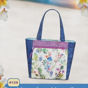 May include: A tote bag with a floral print pocket and blue handles. The bag has a blue and purple trim. The text "Hanalei Handbag" is displayed on the image. The bag is designed by Pink Sand Beach Designs.
