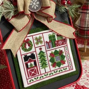 May include: A framed cross-stitch sampler with a red, green, and white color scheme. The sampler features a variety of Christmas-themed designs, including a house, a tree, a wreath, and presents. The frame is red with a green mat and a burlap bow with a red ribbon. The text "Deck the Halls" is printed below the frame.