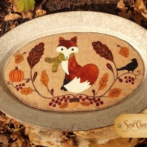 May include: A brown and white fox with a green scarf is the focal point of this autumn-themed needlepoint design. The fox is surrounded by leaves, berries, and a small bird. The design is stitched on a burlap background and is displayed on a metal tray. The text "Sew Cherished" is visible in the lower right corner.