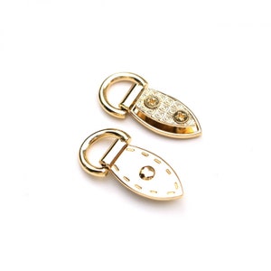 May include: Two gold-toned metal belt loops with a decorative design. The loops have a D-ring on one end and a flat, oval-shaped piece on the other end.