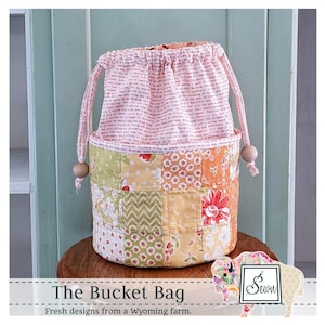 May include: A patchwork bucket bag with a drawstring closure. The bag features a variety of floral and geometric patterned fabric squares in shades of orange, yellow, and green. The top and drawstring are a pink fabric with printed text. The bag has wooden beads.