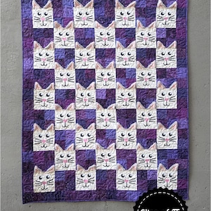 May include: A purple and white quilt with a pattern of white kitten faces. The quilt is titled "Kitten Cuddles" and has a label that says "Slice of Pi Quilts".