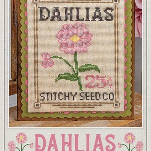 May include: Cross-stitch pattern featuring a pink dahlia flower with a yellow center, the text "DAHLIAS" and "STITCHY SEED CO." on a beige background. The design is framed with green and pink scalloped edges, mounted on a wooden board.