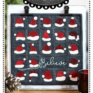 May include: A Christmas-themed embroidery pattern featuring a gray background with 25 red and white Santa hats, with the word "Believe" stitched in white. The finished size is 11 3/4 inches by 11 3/4 inches. The pattern is designed by Lisa Bongean.