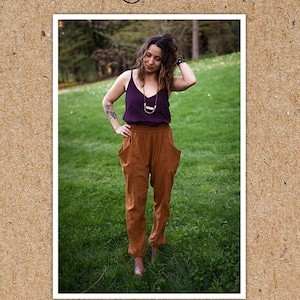 May include: A woman wearing a purple tank top and brown pants with large pockets stands in a grassy field. The pants are called "Arenite Pants" and the image is from the "Sew Liberated" brand.