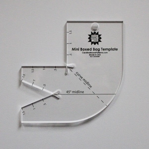 May include: A clear plastic template for making mini boxed bags. The template has markings for a 45 degree midline and a curved midline. The template is labeled "Mini Boxed Bag Template" and includes the website address "CarolinaMoorePatterns.com".