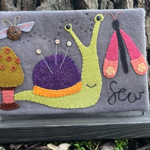 May include: A felt wall hanging with a green snail, purple pincushion, orange and brown mushroom, and a pink butterfly. The snail has a smiling face and the text "Sew" is embroidered on the gray background.
