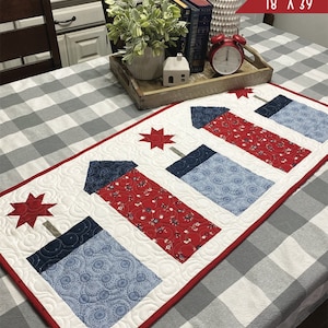 May include: Firework Frenzy table runner, designed by Primrose Cottage, measures 18 inches by 39 inches. The runner features a white background with four red, white, and blue firework designs. The runner is displayed on a gray and white checkered tablecloth.