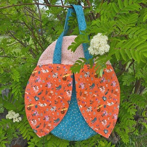 May include: A fabric bag shaped like a ladybug with orange wings featuring a pattern of small fairies and mushrooms, a teal polka-dot underbody, and teal handles. The text #387 FLUTTERBUG BAG and Sew A Story BY JENNIFER LONG Craft Pattern are visible.