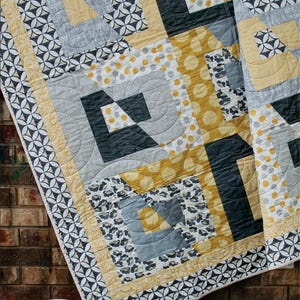 May include: A quilt pattern called "Tabitha" by Swirly Girls. The quilt is made with fat quarters and features a geometric design in shades of gray, yellow, and white. The pattern is fat quarter friendly.