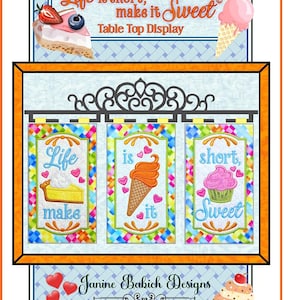 May include: Three colorful embroidered panels with the text "Life is short, make it Sweet" and images of a pie, ice cream cone, and cupcake. The panels are hung on a black metal rod. The text "Janine Bakich Designs" is below the panels.
