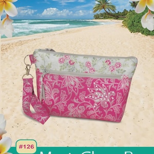 May include: A pink and white floral patterned Maui Glam Bag with a wrist strap. The bag has a zipper closure and a silver-colored metal clasp. The background shows a beach scene with the text "Pink Sand Beach Designs" and "Maui Glam Bag".